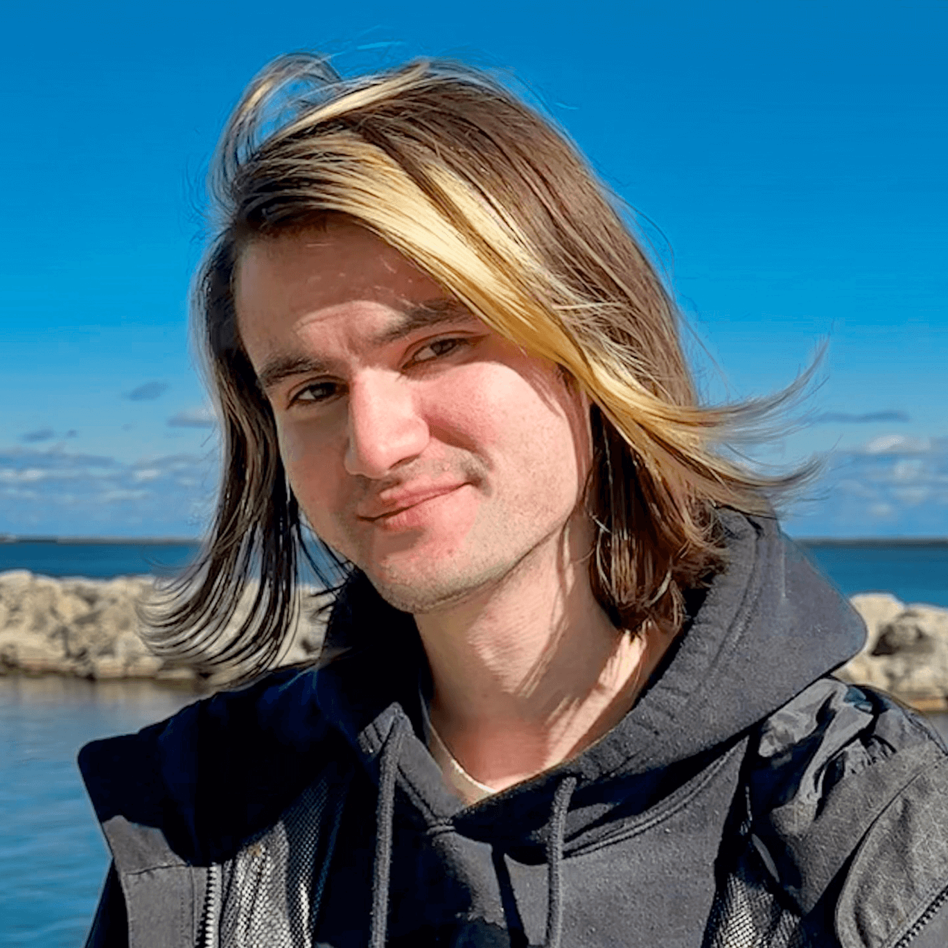 Profile of Daniel Killough, a long haired person slightly smiling at the camera, wearing a black hoodie and standing in front of a lake with rocks.